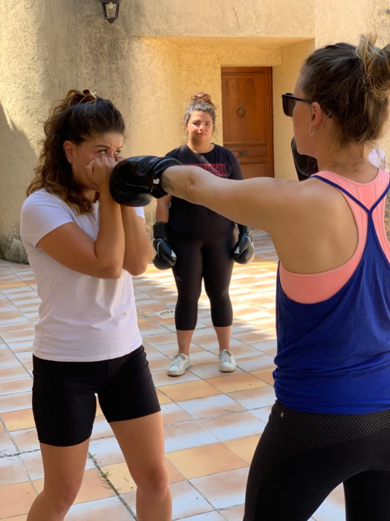 Deux femmes en garde de boxe dans une cour ensoleillée, la coach en arrière-plan